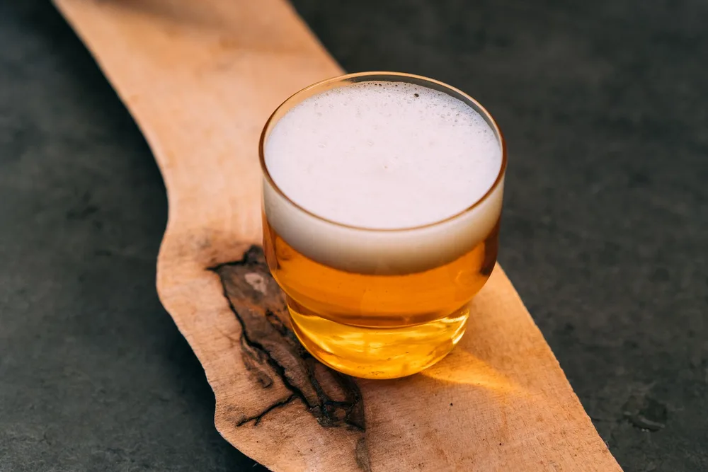 Verre de cidre breton ambré doré avec mousse fine posé sur une planche en bois brut, lumière naturelle