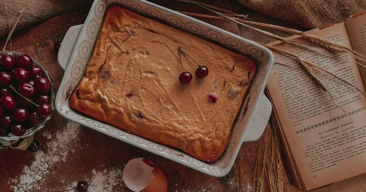 Clafoutis aux cerises encore tiède dans un plat en céramique crème, sur la table en bois de la cuisine de la ferme