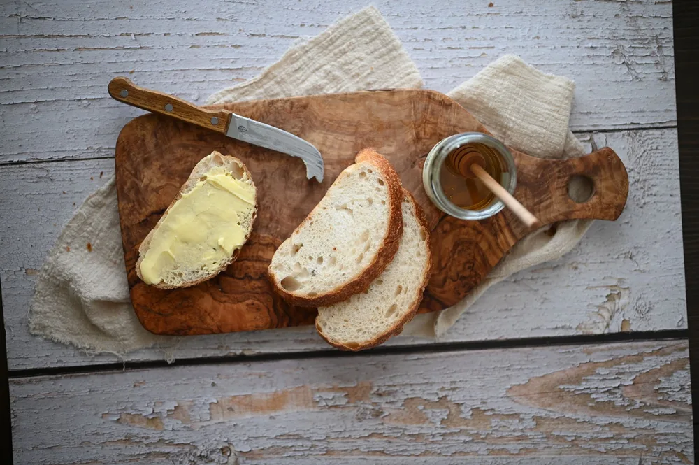 Tranche de pain beurrée sur une planche en bois d'olivier avec un couteau, un torchon en lin et un bocal de miel, vue du dessus