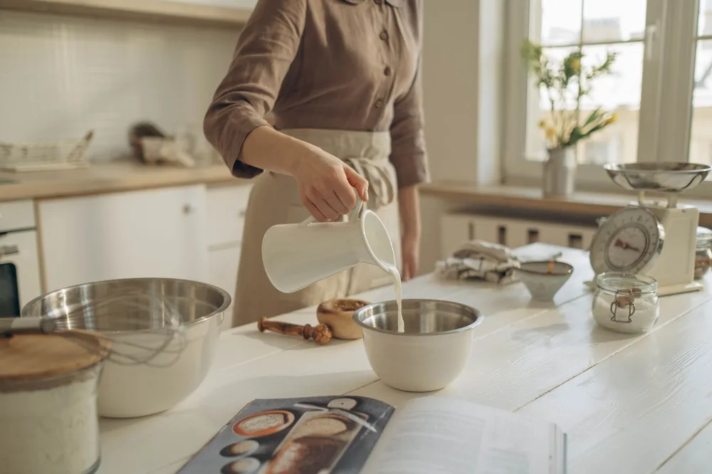 Lait versé lentement depuis un pichet blanc dans un bol en inox, plan de travail en bois, balance et accessoires de cuisine en arrière-plan